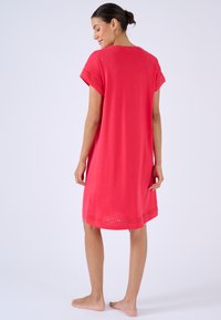 Mujer descalza de pie, vistiendo un vestido rojo de manga corta hasta la rodilla con bordes calados en las mangas y el dobladillo, de espaldas y con la cabeza girada hacia la izquierda.