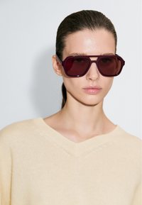 Vrouw met naar achteren gekamde haren, oversized bordeauxrode zonnebril en een crèmekleurige trui met V-hals tegen een effen achtergrond.