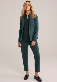 Žena stojící v tmavozeleném saku, odpovídajících kalhotách, tmavozelené blůze a černých mokasínech na prostém béžovém pozadí.