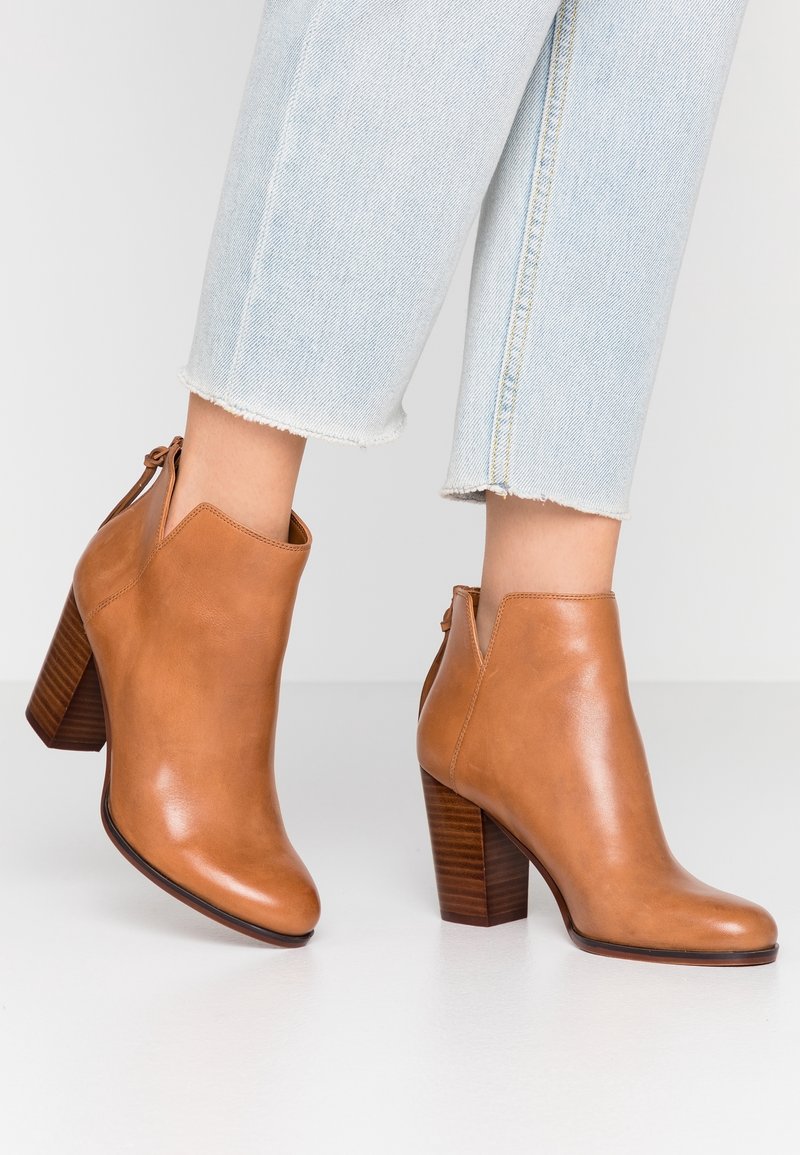 Stivaletti alla caviglia in pelle color cuoio con finitura liscia, punta appuntita e tacco impilato. Chiusura con zip sul retro, abbinati a jeans azzurri con risvolto.