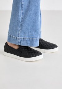 Des chaussures noires à enfiler avec une texture matelassée, une semelle blanche et un orteil arrondi, associées à un jean large bleu clair, coupé au-dessus de la cheville.