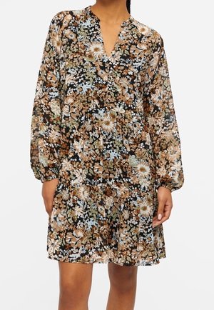 Woman wearing a long-sleeve, knee-length floral dress with a V-neck and loose, gathered cuffs in brown, black, white, and light blue tones.