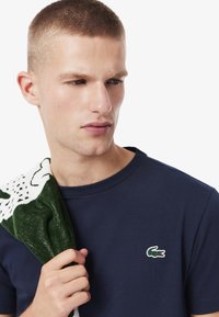 Joven con cabello corto que lleva una camisa azul marino con un logo de cocodrilo verde, sosteniendo un paño de diseño verde y blanco sobre su hombro.