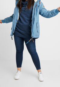 Giacca blu con motivo e cappuccio foderato in pelliccia, indossata sopra una felpa blu scuro. Abbinata a jeans scuri e sneakers bianche.