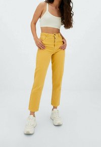 Gula högmidjade jeans med rak ben design och femfickorsstil, kombinerade med en ljusfärgad, ribbad crop top och vita sneakers.