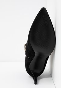 Chaussure à talon haut en daim noir avec un bout pointu et un talon stiletto fin. Elle présente un détail accent doré et une semelle extérieure noire texturée.