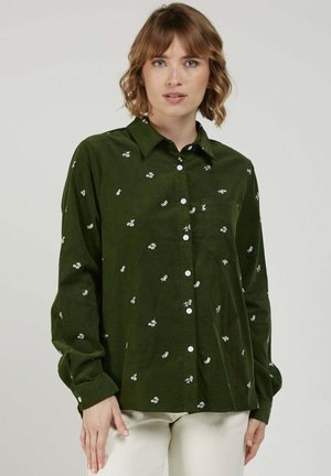 Femme aux cheveux courts et ondulés portant une chemise boutonnée vert foncé avec de petits motifs floraux blancs et un pantalon clair.