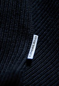 Schwarzer Strickstoff mit gerippter Textur, versehen mit einem weißen Etikett mit der Aufschrift "LINDENBERGH", das am Rand angenäht ist.