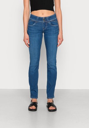 Jeans skinny de denim azul con dos bolsillos frontales, una cintura de altura media y costuras en contraste. Combinados con sandalias negras. Textura suave.