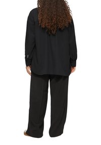 Zwarte button-up shirt met lange mouwen, ontspannen pasvorm, textuurstof, en zwarte wijde broek, zichtbaar aan de onderkant van de afbeelding.