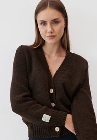 Brązowy dziergany kardigan z dekoltem w serek, sześcioma drewnianymi guzikami i prążkowanymi mankietami. Zawiera metkę z materiału na lewym rękawie.