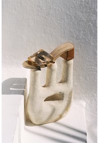 Sandal con tacco a blocco in legno di tonalità dorata, con fasce in rete, esposta su un piedistallo scultoreo a forma di mano.