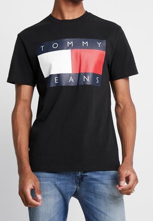 Mann trägt ein schwarzes T-Shirt mit kurzen Ärmeln und großem Tommy Jeans Logo in Weiß, Rot und Marineblau, kombiniert mit hellblauen Jeans.