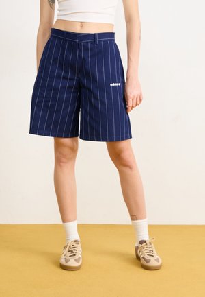 Persoon die marineblauwe Adidas shorts met fijne strepen draagt, een wit cropped topje, witte sokken en beige sneakers, staand op een mosterdkleurige vloer.