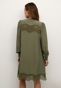 Olīvu zaļa kleita ar garām pūkainām piedurknēm, ar dekoratīvu mežģīņu apdari pie kakla un apakšmalas, kā arī gludu, plūstošu auduma tekstūru.