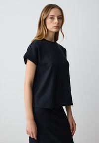 Top nero a maniche corte con vestibilità ampia e scollatura rotonda. Realizzato in tessuto liscio, presenta maniche arrotolate e un design minimalista.