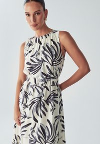 Ramnica brez rokavov z nabranim izrezom, ki prikazuje črne in sive listnate vzorce na svetlem bež ozadju. Elastični pas, tekoča tkanina.