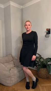 Czarna sukienka z długim rękawem i rozkloszowanym dołem, noszona z białymi skarpetkami i czarnymi butami. W tle widoczna beżowa kanapa w paski oraz zielona roślina w plecionym koszu.