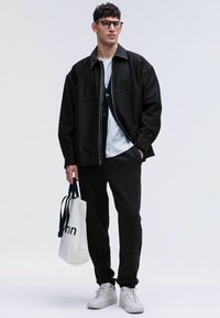 Schwarze Zip-Jacke über einem weißen T-Shirt, schwarze Hose und weiße Sneakers. Hält eine Canvas-Tasche mit einem bedruckten Logo. Lockige Haare und Brille.