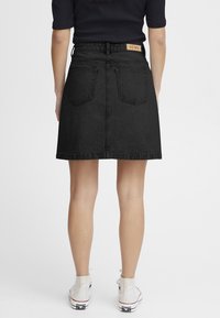 Jupe en denim noir à coupe droite, dotée d'une taille haute, de deux poches arrière et d'une étiquette en cuir à la taille. Portée avec des baskets blanches.