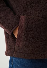 Brown fleece jacket with a soft, textured surface, featuring a ribbed accent near the pocket and hem. Casual fit with a comfortable look.