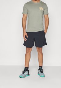 Camisa de manga corta verde oliva con un gráfico, combinada con pantalones cortos deportivos negros. Zapatillas gris y negras con acentos turquesa.