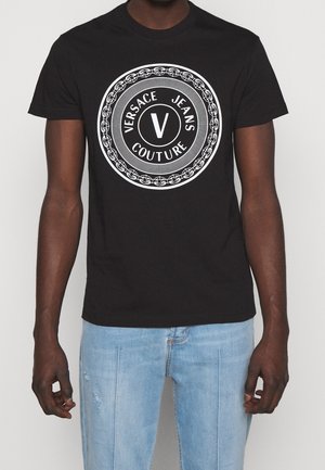 Czarna koszulka z białym, okrągłym logo Versace Jeans Couture na klatce piersiowej, noszona z jasnoniebieskimi dżinsami, pokazana od szyi do bioder.