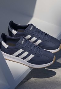 Zapatillas Adidas azul marino con franjas blancas y suelas de goma descansando sobre una superficie geométrica blanca con luz suave.