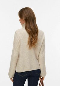 Bež pulover z zvončnim ovratnikom in rebrastim robom ter dolgimi rokavi, kombiniran s temnimi kavbojkami. Material je videti mehak in teksturiran.