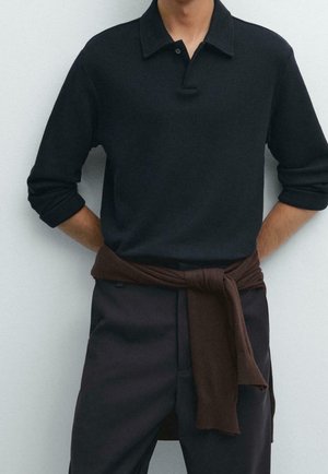 Person iført en sort langærmet polo med opkrogede ærmer og en brun sweater bundet rundt om taljen mod en ensfarvet baggrund.