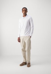 Cămașă polo albă cu mâneci lungi și un mic logo, asortată cu pantaloni bej deschis și adidași din suede maro. Design simplu și curat.