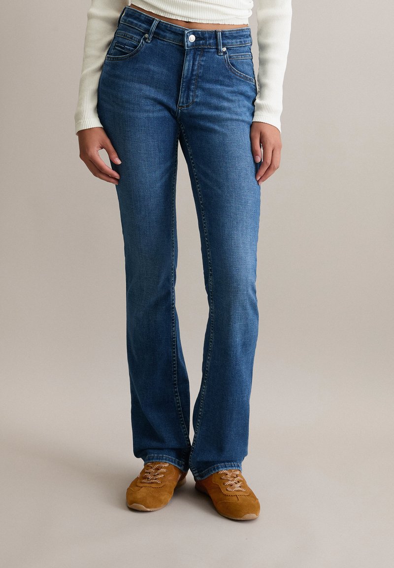 Blaue Denim-Jeans im Straight-Leg-Schnitt mit fünf Taschen, einem Knopfverschluss und dezenten Nähdetails, getragen mit braunen Schnürschuhen.