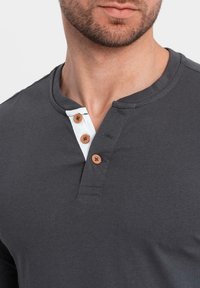 Graues langärmliges Henley-Shirt mit rundem Ausschnitt, das drei Holzknöpfe und ein kontrastierendes weißes Blenden-Detail aufweist.