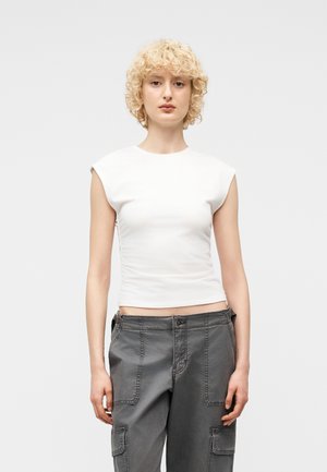 Ung person med krøllet blondt hår iført en hvid, ærmeløs tætsiddende top og løstsiddende mørkegrå cargobukser mod en ensfarvet baggrund.