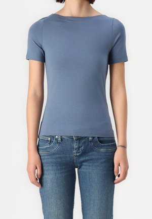 Persona que lleva una camiseta azul de manga corta ajustada y vaqueros azules, de pie frente a un fondo blanco liso.