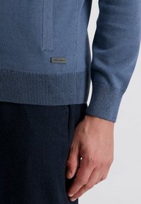 Blauer Strickpullover mit strukturierten Rippbündchen und -saum, ausgestattet mit einem kleinen schwarzen Logo-Platinen. Nahaufnahme einer Hand, die auf einem dunklen Stoff ruht.