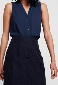 Blusa senza maniche color navy con scollo a V e due bottoni dorati sul davanti, abbinata a una gonna in pizzo color navy con un motivo floreale.