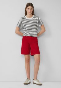 Shorts en coton rouges avec des manches retroussées, assortis à un t-shirt rayé noir et blanc. La tenue est complétée par des baskets beiges et brunes.