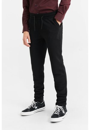 Persoon die zwarte, gestructureerde broek met trekkoord, zwart-witte sneakers en een bordeauxrode blouse met patroon draagt, staand met één hand in de zak.