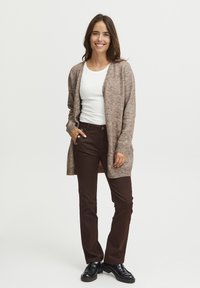 Gilet en tricot marron porté par-dessus un haut blanc, associé à un pantalon marron foncé et des chaussures noires. Le gilet présente une coupe ample et des poignets côtelés.
