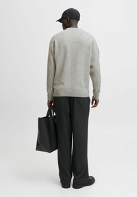 Grijze wollen trui met geribbelde rand, zwarte pet en donkere broek. Zwarte tote bag in de rechterhand. Eenvormige achtergrond.