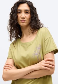 Γυναίκα με κατσαρά μαλλιά που φοράει ένα ανοιχτό πράσινο μπλουζάκι με μοτίβο από μοβ φοινικόδεντρο, με σταυρωμένα τα χέρια, μπροστά σε απλό ανοιχτό υπόβαθρο.