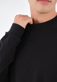 Svart långärmad tröja med rund halsringning, ribbade manschetter och mjuk, slät textur, delvis synlig på en persons överkropp.
