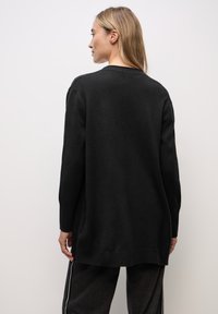 Svart oversized kofta med långa ärmar, ribbad fåll och mjuk textur. Har rund halsringning och avslappnad passform, lämplig för lager-på-lager-stil.