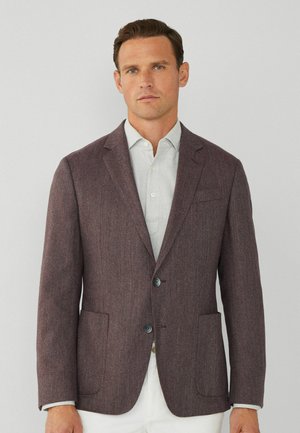 Blazer en chevrons bordeaux avec revers pointus, deux poches avant et deux boutons. Porté par-dessus une chemise à carreaux claire.