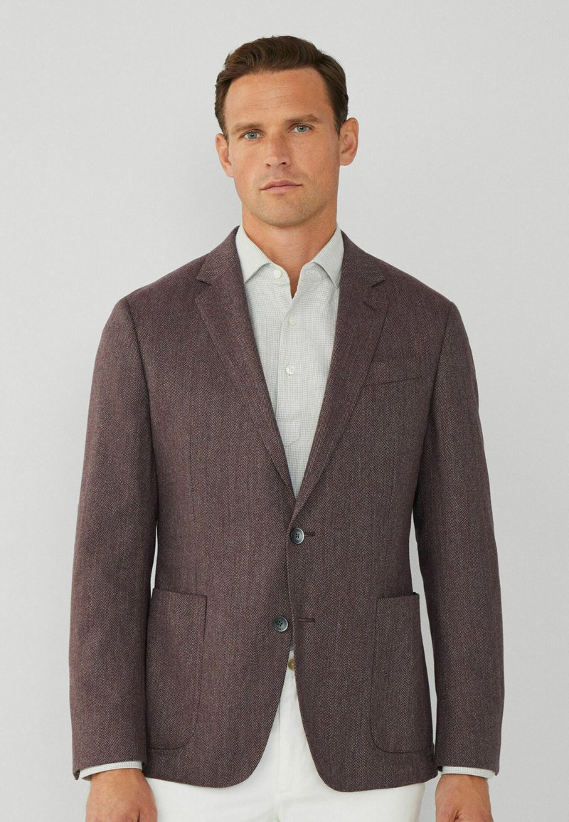 Blazer in tessuto a zigzag bordeaux con rever a punta, due tasche anteriori e due bottoni. Indossato sopra una camicia a quadratini chiara.