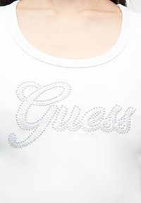 Gros plan sur un haut blanc à côtes avec un col dégagé, orné du mot « Guess » épelé en petits clous brillants sur la poitrine.