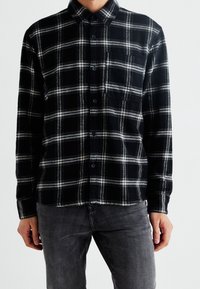 Svart och vit rutig flanellskjorta med knapp-ner krage, framficka och långa ärmar. Tyget har en mjuk textur.