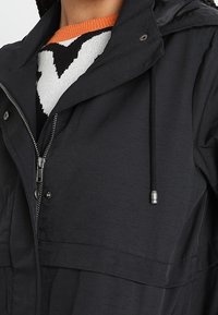 Veste à capuche noire à fermeture éclair avec cordons, portée par-dessus un pull blanc et noir avec un col orange.