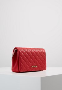 Červená prošívaná kabelka z umělé kůže s řetízkem, která má na přední straně zlatý logový akcent a obdélníkový tvar.